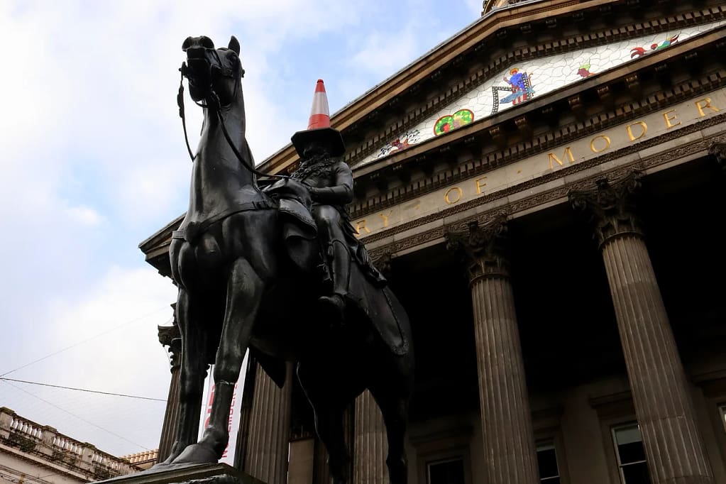 Photograph of an equetrian statue in front of an official building with columns, with a traffic cone on the head of the rider.