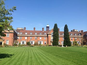 View of St. Hugh's College from the gardens, showing a vibrant green lawn leading up to a red brick stone building.