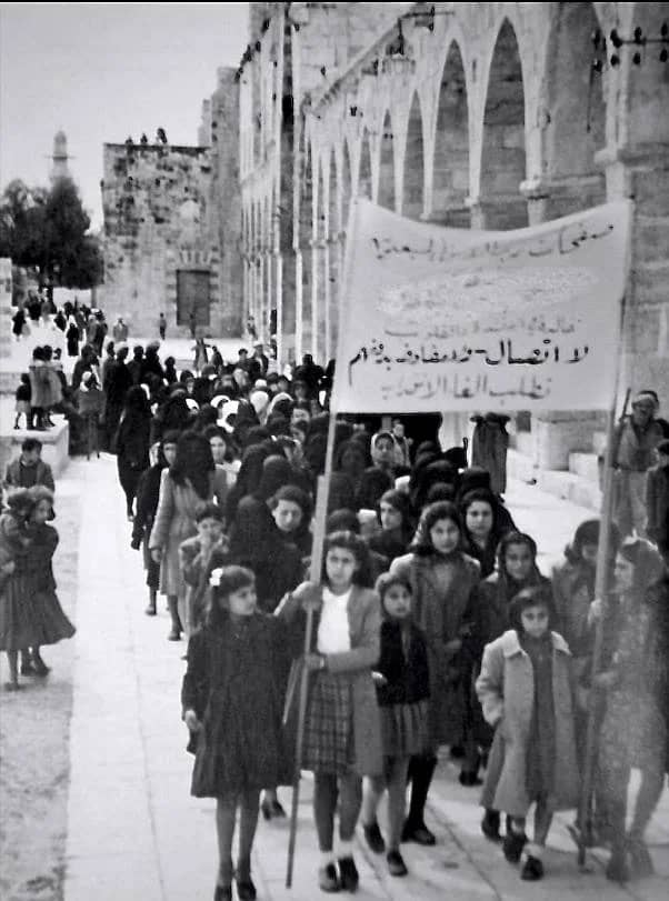 Black and white picture of women and girls protesting, holding a banner with arabic script.