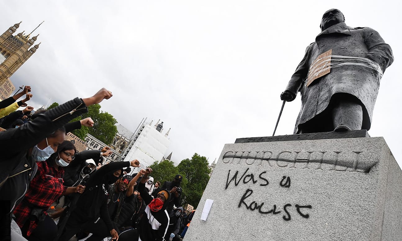 Photograph of a statue surrounded by protesters raising a fist, with graffiti on the base.