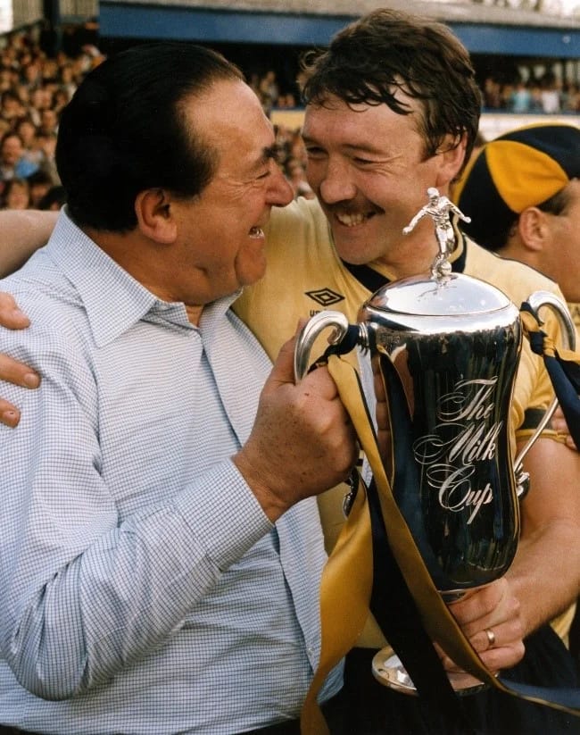 Photograph of two men embracing while holding a silver football cup. 